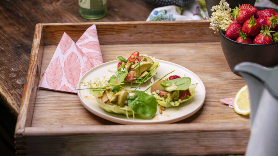 Auf einem Holztablar steht ein Teller mit einem Avocado-Erdbeer-Salat, sommerlich frisch angerichtet