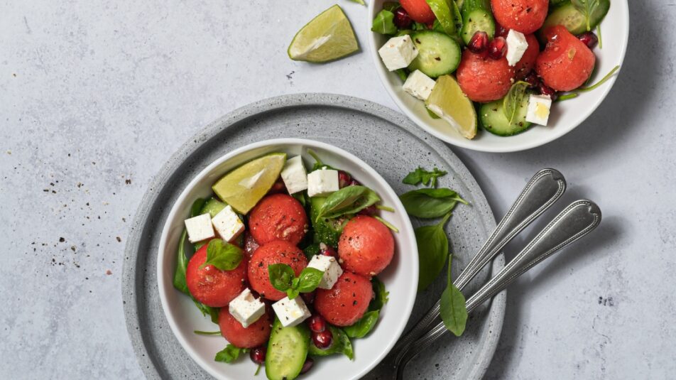 Drei Schüsseln mit sommerlichem Salat aus Wassermelone, Gurke, Feta, Granatapfel und frischen Kräutern, garniert mit Limettenstücken auf hellem Untergrund