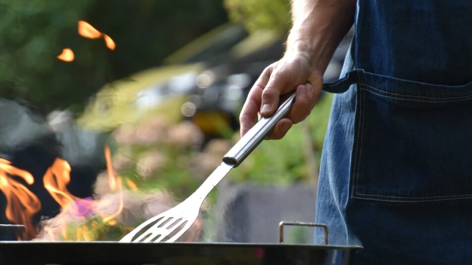 Grillszene im Garten mit einer Person in Schürze, die eine Grillzange über einem offenen Holzkohlegrill mit lodernder Flamme hält