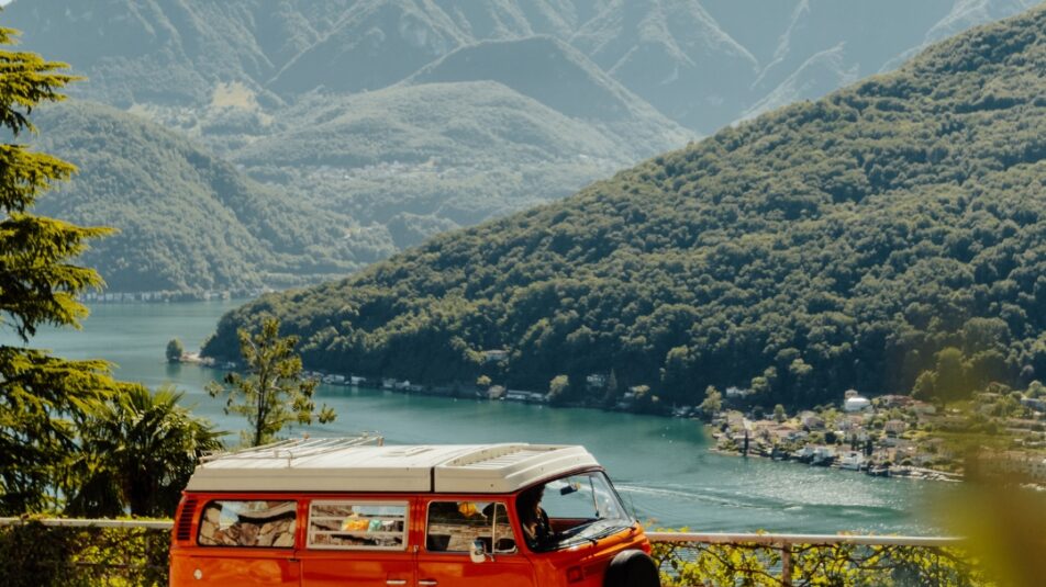 Orangefarbener VW-Bus fährt auf einer Strasse mit Blick auf einen See und umliegende grüne Berge in der Schweiz