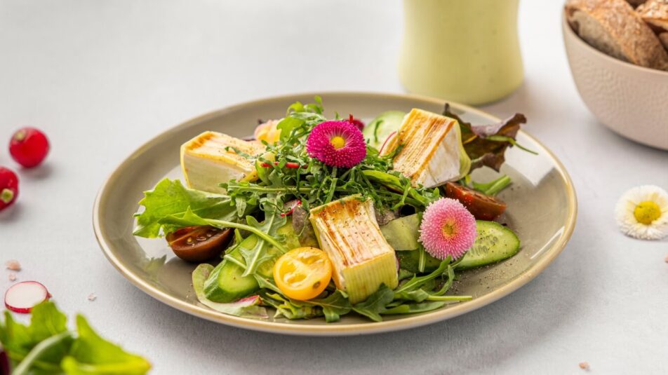 Teller mit frühlingshaftem Salat aus Blattsalat, Brie in Lauch eingewickelt und essbaren Blumen, daneben eine Flasche Brunos Salatsauce Bärlauch