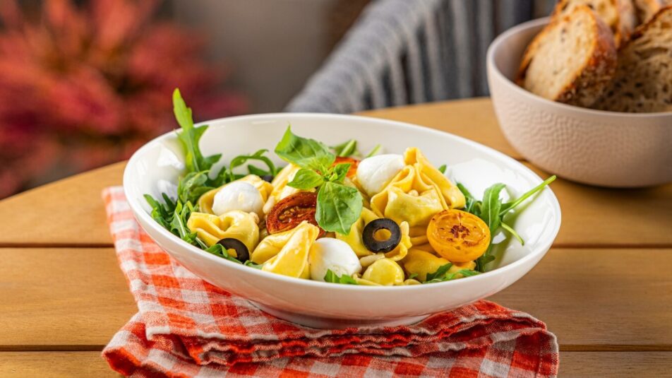 Tortellinisalat mit Cherry-Tomaten und Mozzarella auf einem Terrassentisch. Im Hintergrund eine Pfeffermühle und Brot.