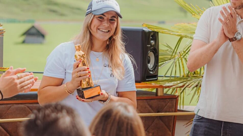 Delia Durrer, eine Skifahrerin, erhält bei der Brunos Talents Challenge eine goldene Salatsauce als Pokal. Sie steht lächelnd in der Mitte des Bildes und hält die goldene Salatsauce von Brunos in der Hand. Um sie herum stehen Personen, die applaudieren. Im Hintergrund ist eine grüne Landschaft zu sehen.