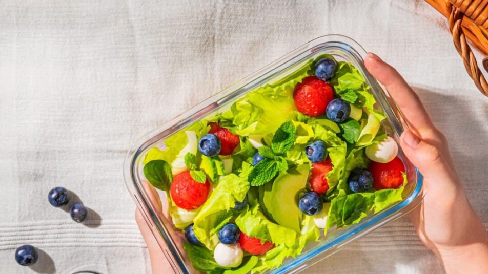 Ein rechteckiger Glasbehaelter mit einem frischen Salat aus gruenen Blaettern, Wassermelonenbaellchen, Blaubeeren, kleinen Mozzarellakugeln und Minzblaettern. Der Behaelter wird von zwei Haenden gehalten. Im Hintergrund ist ein weisses Tuch und ein Picknickkorb zu sehen, daneben einige verstreute Blaubeeren und Minzblaetter.