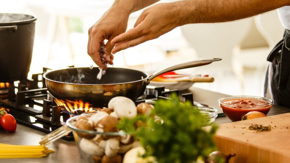 Person bereitet ein Gericht auf einem Herd zu, umgeben von frischen Zutaten wie Tomaten, Paprika und Pilzen – repräsentiert die praktischen Tipps der 10 Küchen-Lifehacks.